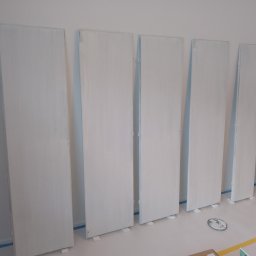 USŁUGI BUDOWLANE Krzysztof Gołębiowski - Białe, świeżo pomalowane skrzydła drzwiowe o prostych kształtach, ustawione rzędem w pomieszczeniu, zabezpieczone taśmą malarską przy podłodze. Widoczne ślady farby.