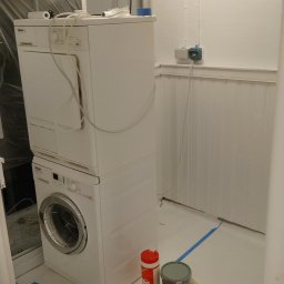 USŁUGI BUDOWLANE Krzysztof Gołębiowski - Białe pomieszczenie w trakcie malowania: zaklejona podłoga, puszki z farbą, maszyna do prania i suszenia. Widoczne białe panele ścienne i rury pod sufitem.