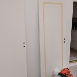 USŁUGI BUDOWLANE Krzysztof Gołębiowski - Białe drzwi z zamontowanymi klamkami, jedne z surową ramą. Na podłodze narzędzia, w tle widok na wnętrze pomieszczenia. Prace wykończeniowe w trakcie.