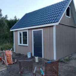 Mały, parterowy budynek z ciemnym dachem i beżową elewacją z pionowych paneli. Widoczne okno, drzwi wejściowe, worki z materiałami budowlanymi i dwa krzesła.