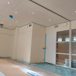 Wnętrze w trakcie układania paneli podłogowych. Podłoga częściowo zakryta folią ochronną i kartonem. Ściany pomalowane, zabezpieczone taśmą malarską. Widoczne puszki paneli.