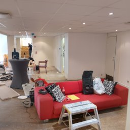 USŁUGI BUDOWLANE Krzysztof Gołębiowski - Pokój w trakcie remontu: czerwona sofa z poduszkami, drabina, narzędzia i worki budowlane na podłodze. W tle widoczna osoba na schodach. Oświetlenie sufitowe.