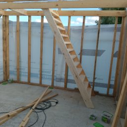 USŁUGI BUDOWLANE Krzysztof Gołębiowski - Wnętrze szkieletu budynku w trakcie budowy. Widoczne drewniane elementy konstrukcyjne, schody tymczasowe, folia na ścianach i narzędzia na betonowej podłodze.