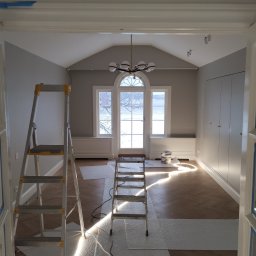 USŁUGI BUDOWLANE Krzysztof Gołębiowski - Pokój w trakcie remontu. Widoczne drabiny, zabezpieczona podłoga i ściany przygotowane do malowania. W tle okno z widokiem na zewnątrz i nowoczesny żyrandol.