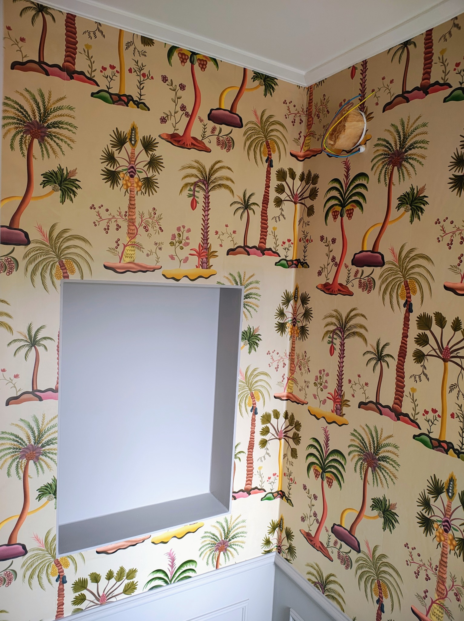 Ściana z tropikalną tapetą w odcieniach beżu i zieleni, z wnęką pomalowaną na szaro. Widoczne kable wystające ze ściany. Dekoracyjne wykończenie.