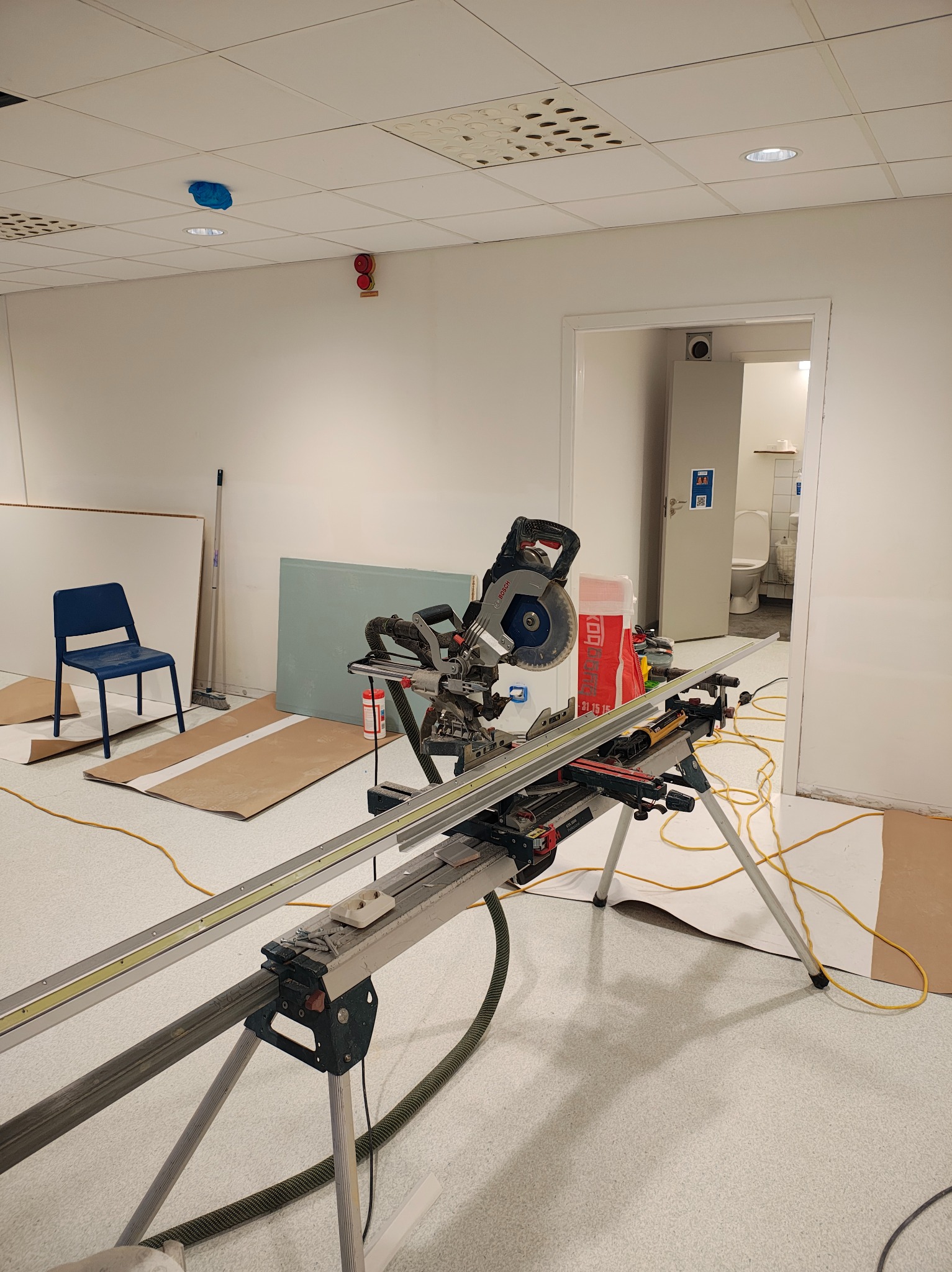 Piła ukośnica na statywie w pomieszczeniu z płytą karton-gips i krzesłem. Widoczny sufit podwieszany i drzwi do toalety. Przygotowania do pracy.