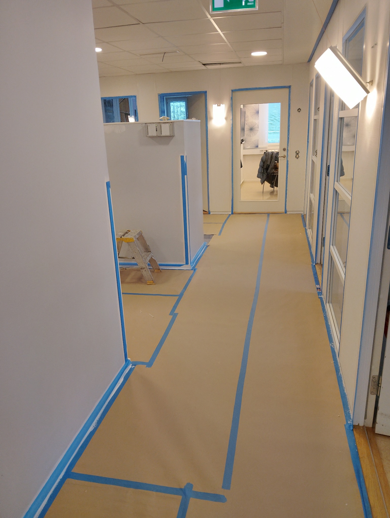 Przygotowanie do malowania: zabezpieczone taśmą i papierem ściany, podłoga w korytarzu biurowym. Widoczna drabina i białe ściany.