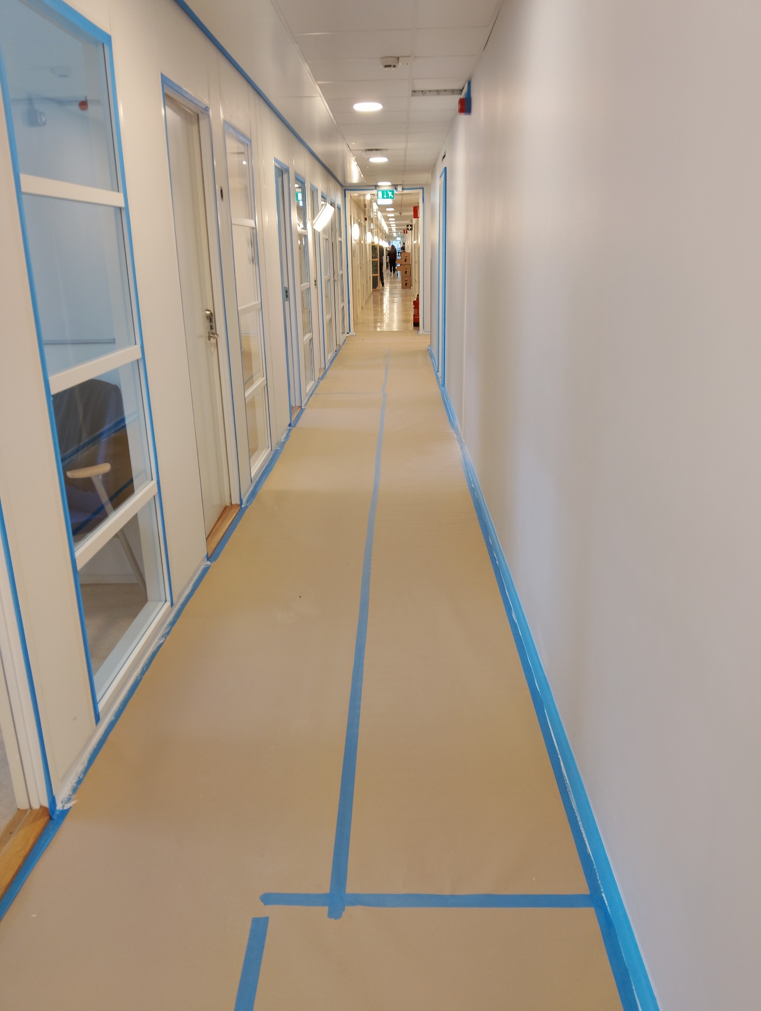 Korytarz zabezpieczony taśmą malarską przed pracami wykończeniowymi. Widoczne białe ściany i podłoga w kolorze beżowym. W tle osoba przechodząca korytarzem.