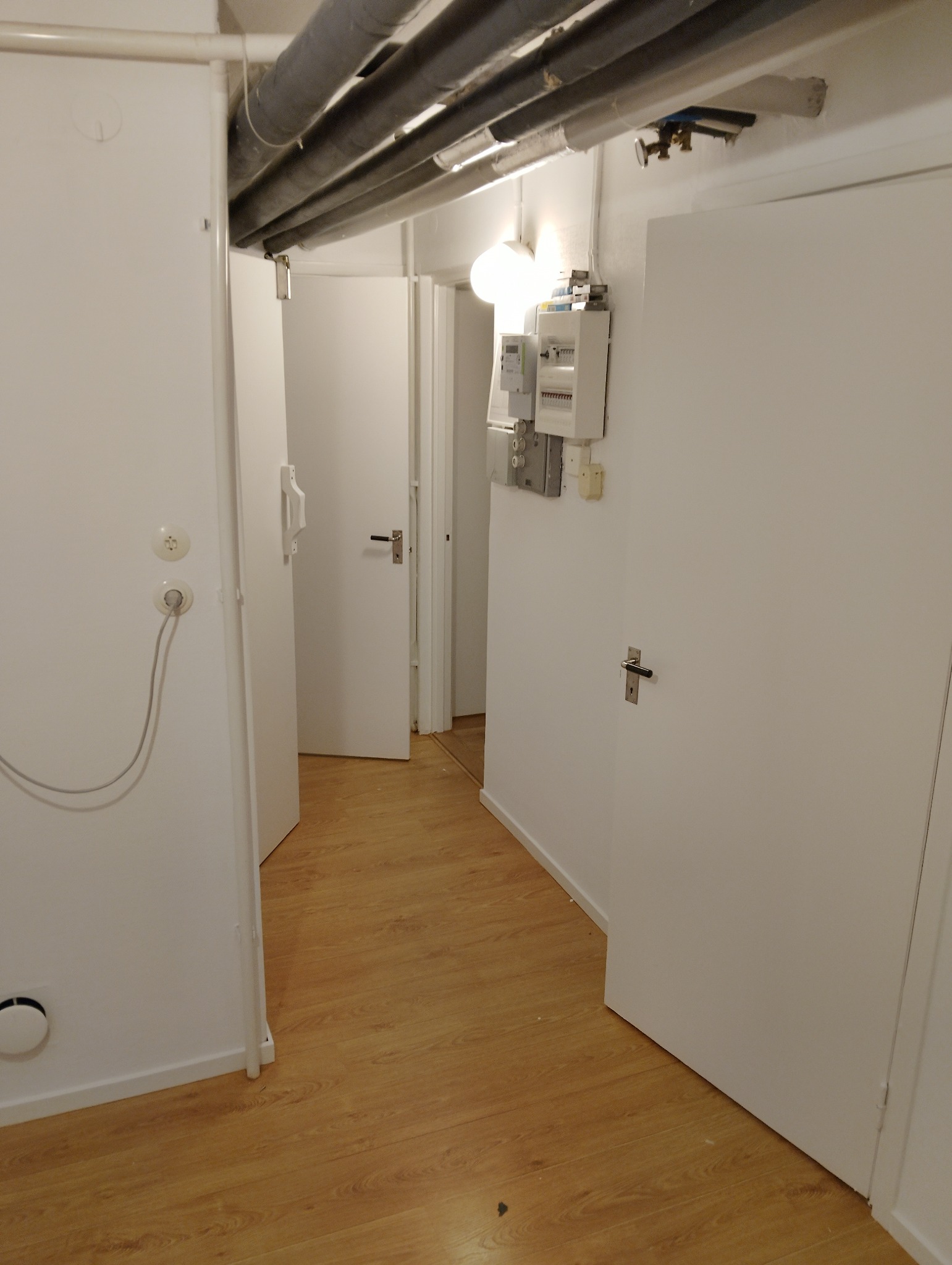 Białe, odświeżone wnętrze korytarza z drewnianą podłogą, widoczne drzwi i instalacje pod sufitem. Minimalistyczny design z elementami technicznymi.