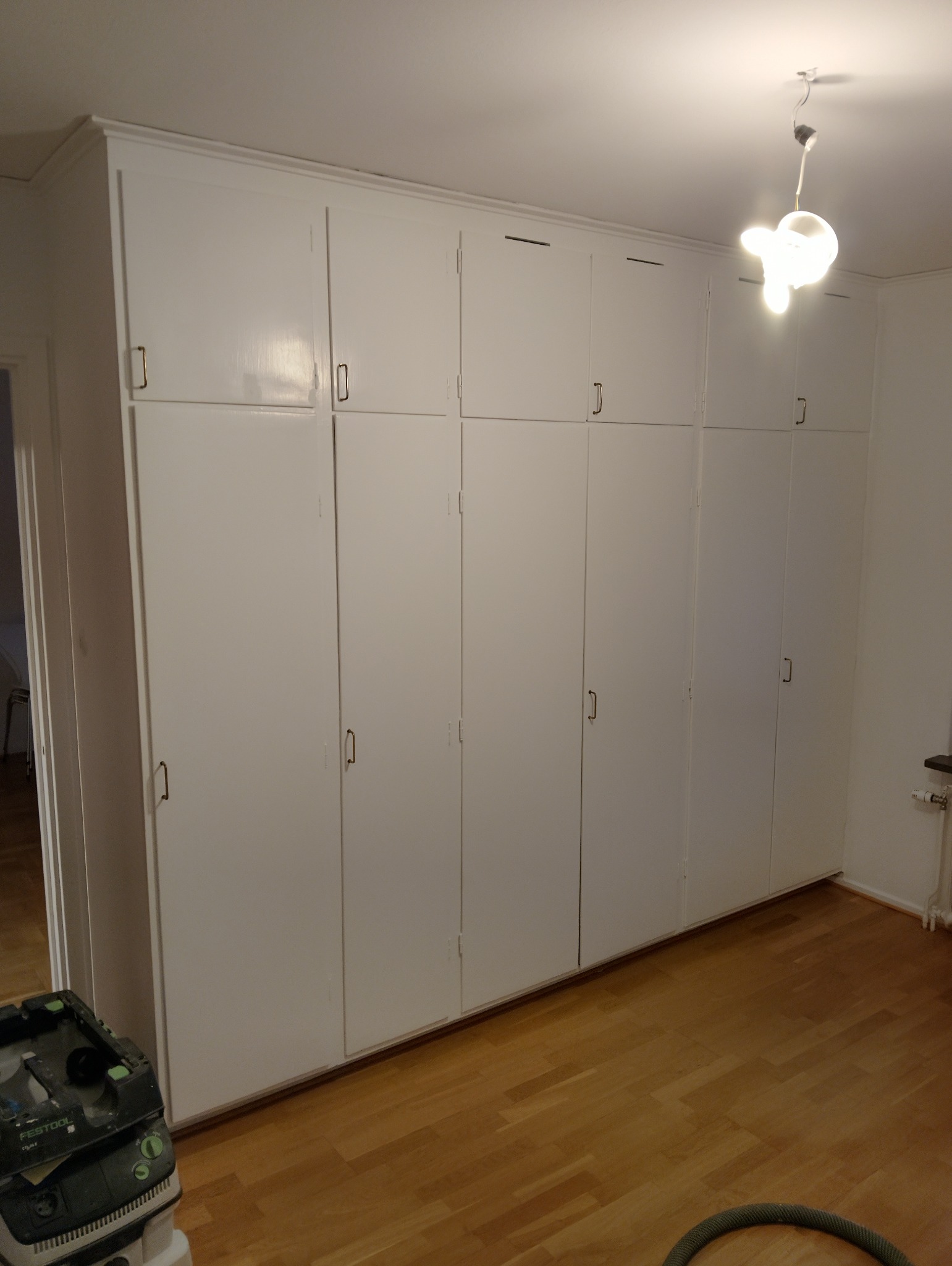 Biała zabudowa szaf wnękowych z drewnianą podłogą. Widoczny sprzęt czyszczący i oświetlenie sufitowe. Realizacja wykończenia wnętrz.