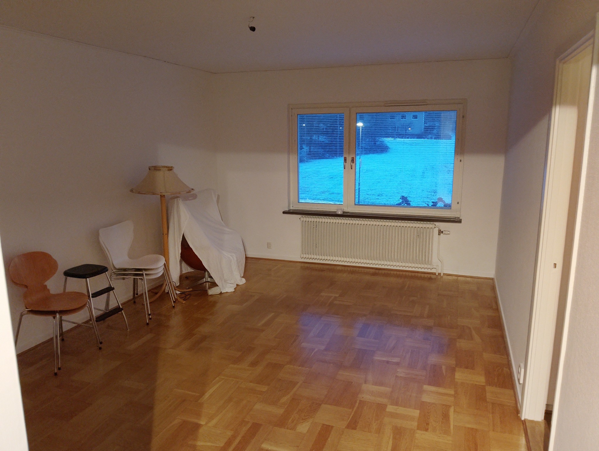 Pusty pokój z parkietem i grzejnikiem w Starogardzie Gdańskim. Widoczne okno z żaluzjami, krzesła i lampa pod abażurem. Przygotowanie do prac wykończeniowych.