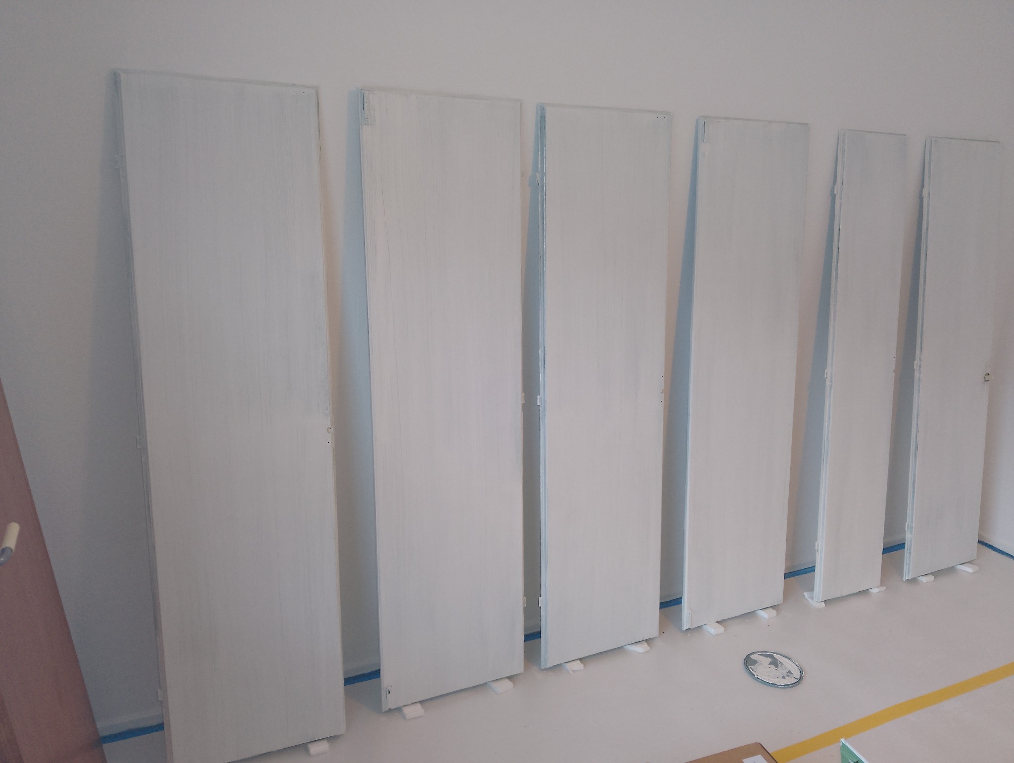 Białe, świeżo pomalowane skrzydła drzwiowe o prostych kształtach, ustawione rzędem w pomieszczeniu, zabezpieczone taśmą malarską przy podłodze. Widoczne ślady farby.