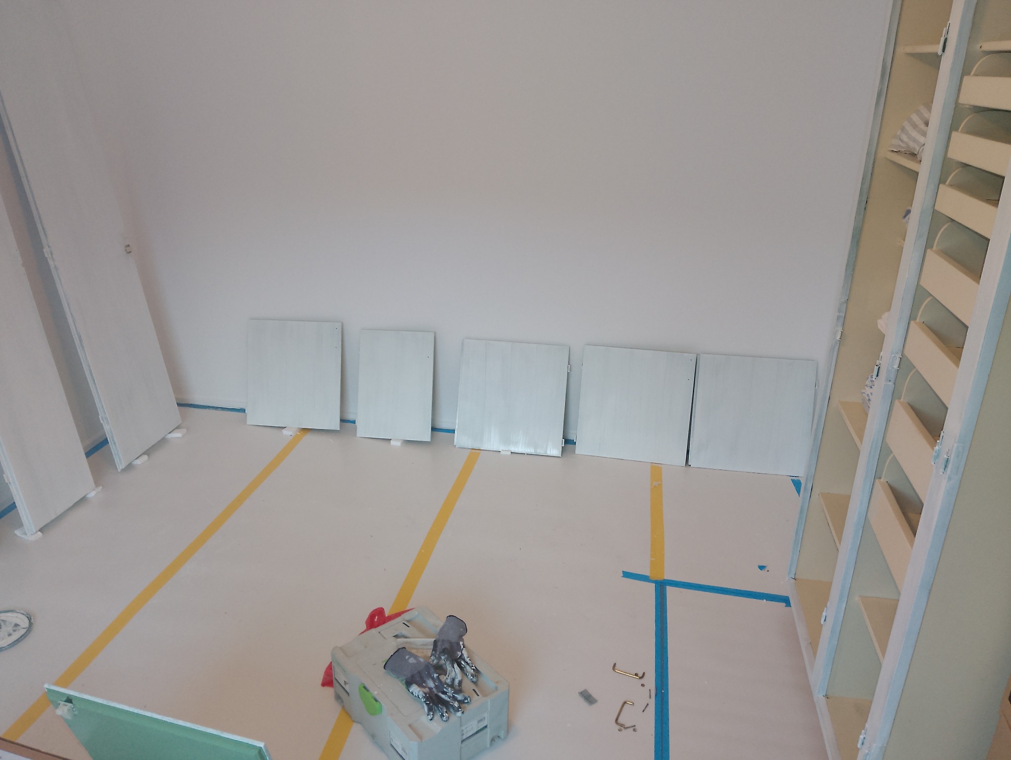 Przygotowane płyty do malowania w pomieszczeniu z zabezpieczoną podłogą. Widoczny kufer z narzędziami i malowane regały.