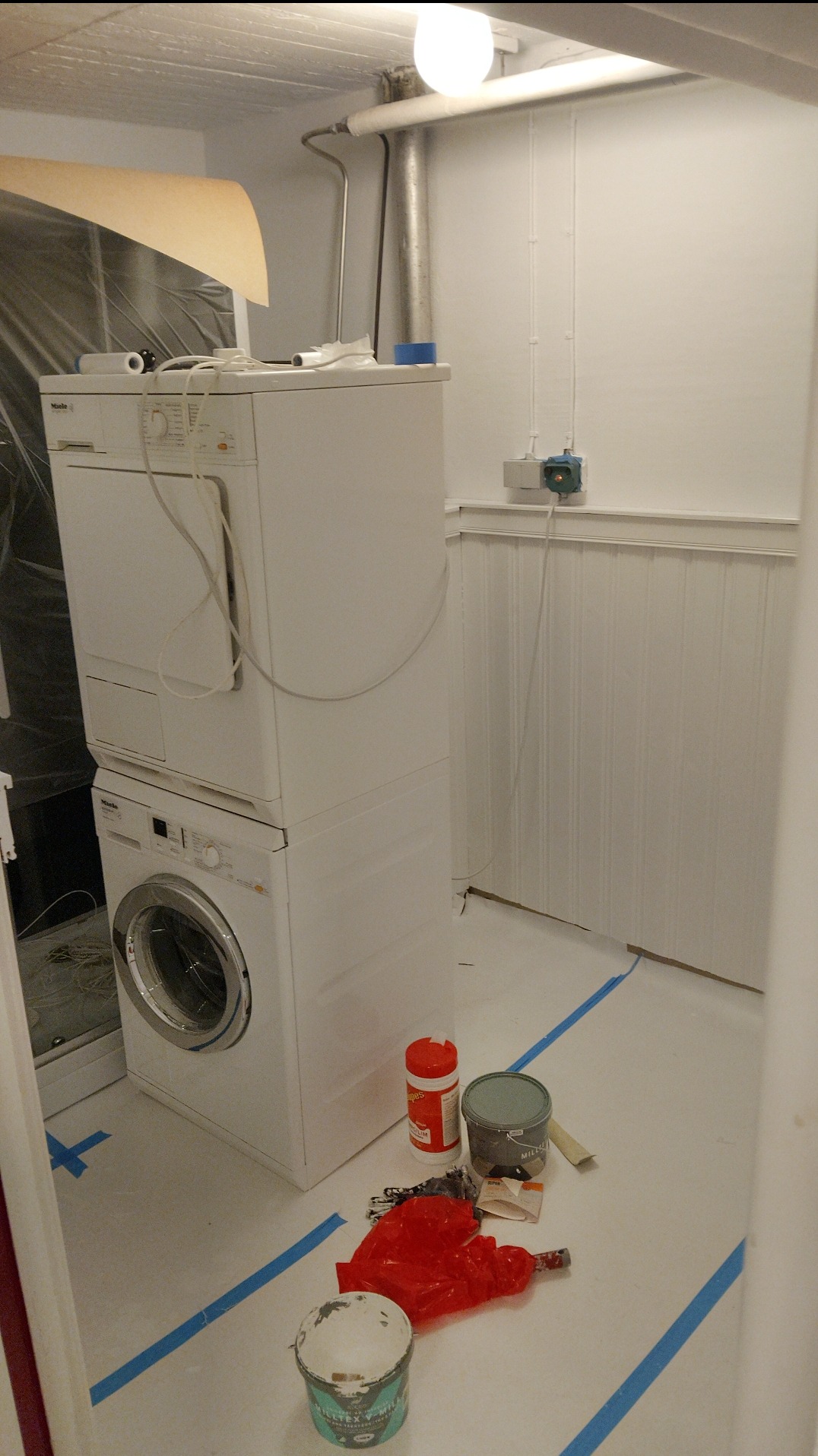 Białe pomieszczenie w trakcie malowania: zaklejona podłoga, puszki z farbą, maszyna do prania i suszenia. Widoczne białe panele ścienne i rury pod sufitem.