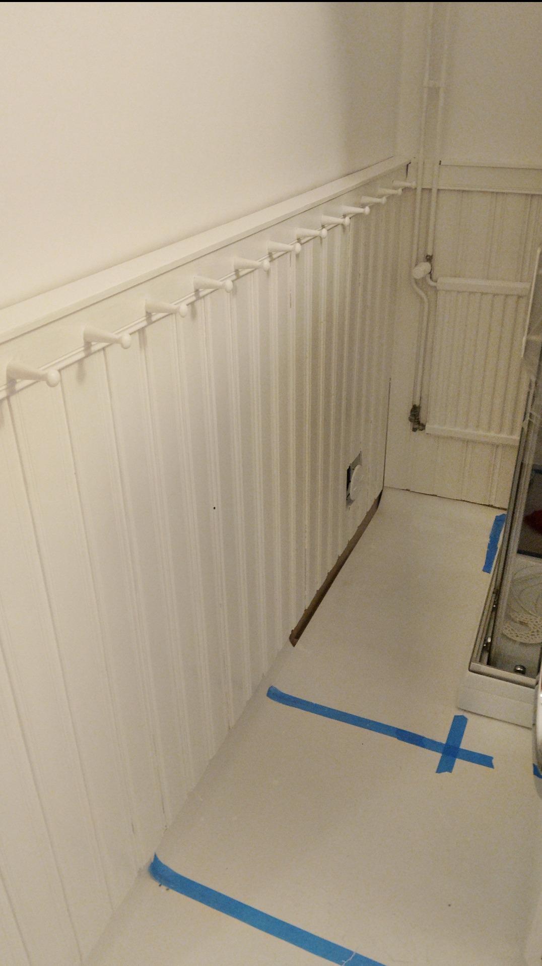 Białe, pionowe panele ścienne z wieszakami i listwą przypodłogową, zabezpieczone niebieską taśmą malarską na podłodze. Widoczny fragment rury.
