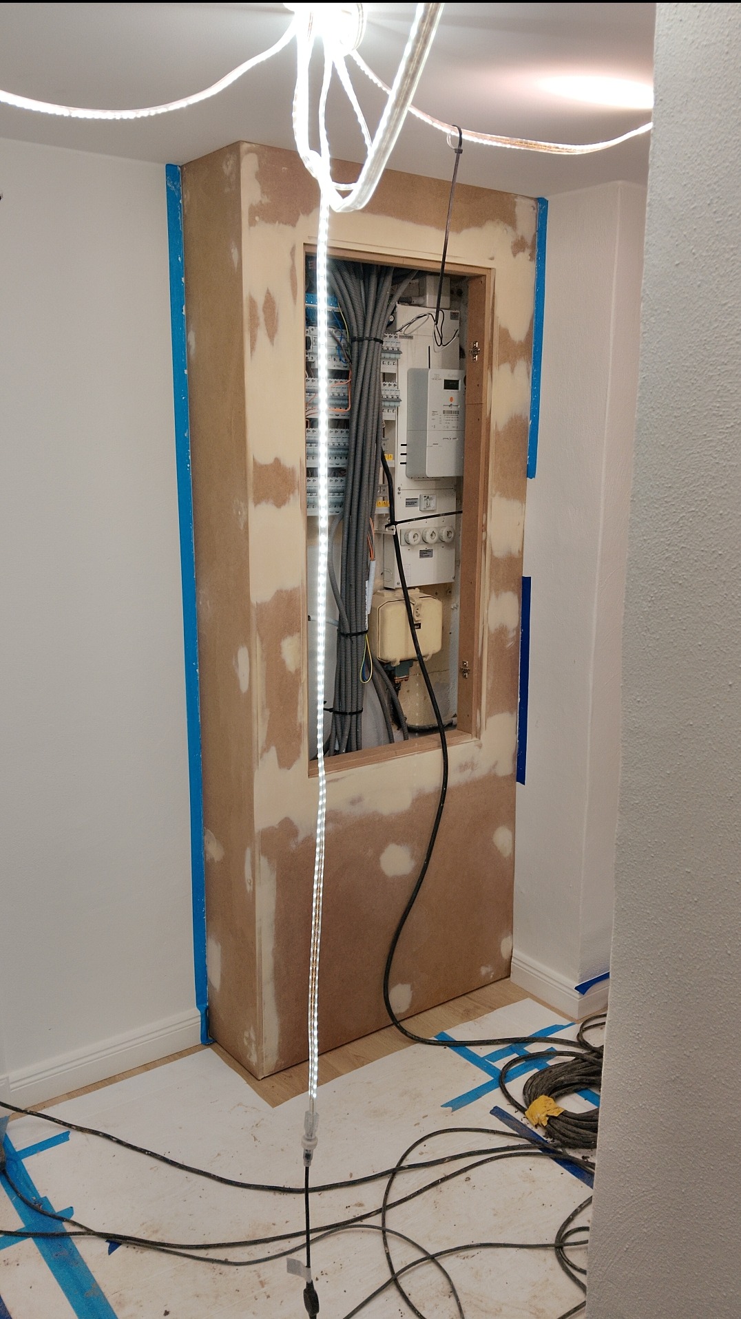 Obudowa szafki elektrycznej w trakcie prac wykończeniowych. Widoczne zabezpieczenia taśmą malarską, przygotowana płyta MDF i przewody elektryczne.