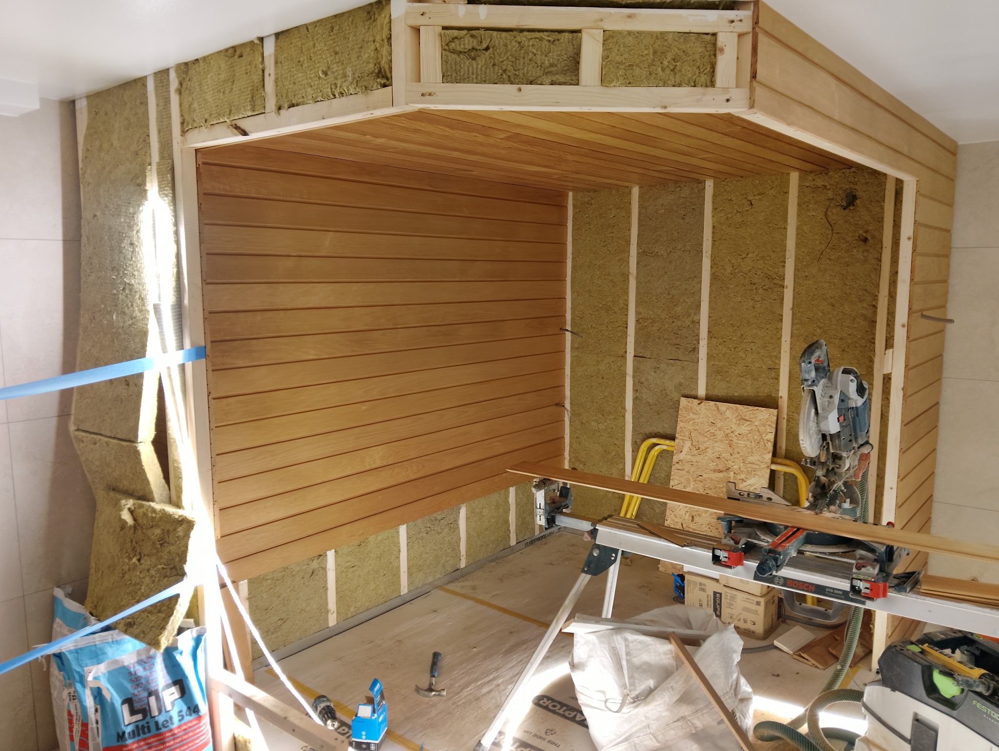 Wnętrze w trakcie prac: montaż drewnianej boazerii na ścianie, ocieplenie wełną mineralną, widoczne narzędzia i materiały budowlane. Kątowe ujęcie z bliska.