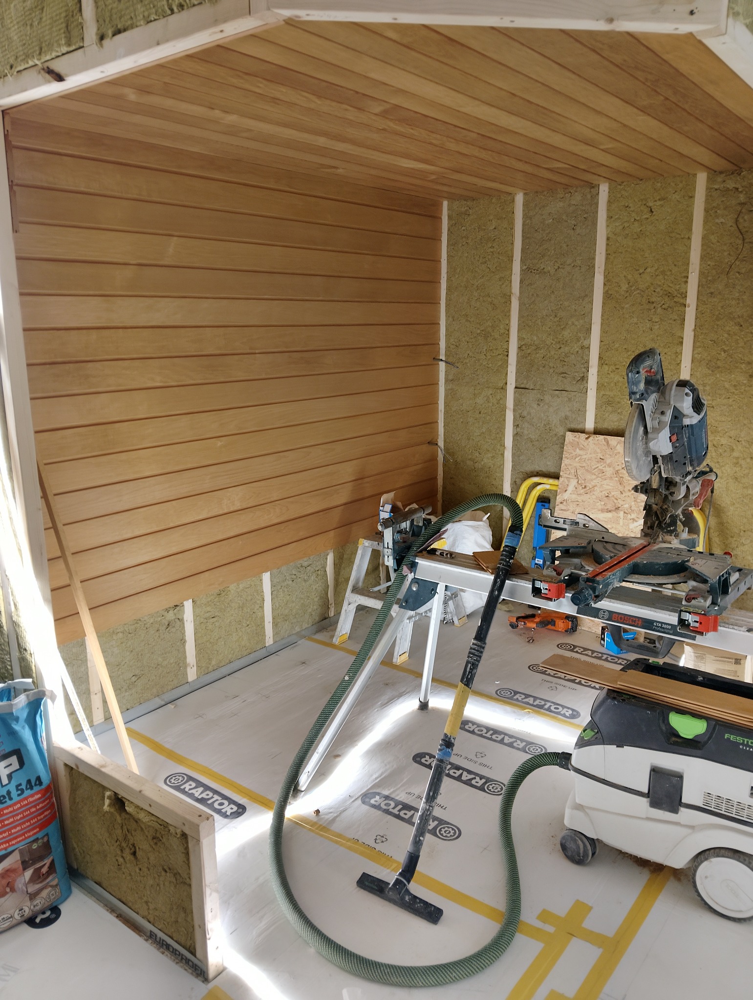 Wnętrze w trakcie wykańczania drewnem, widoczne narzędzia i materiały izolacyjne. Ściany częściowo obłożone boazerią, sufit wykończony drewnem.