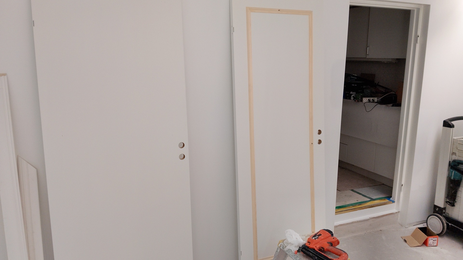 Białe drzwi z zamontowanymi klamkami, jedne z surową ramą. Na podłodze narzędzia, w tle widok na wnętrze pomieszczenia. Prace wykończeniowe w trakcie.