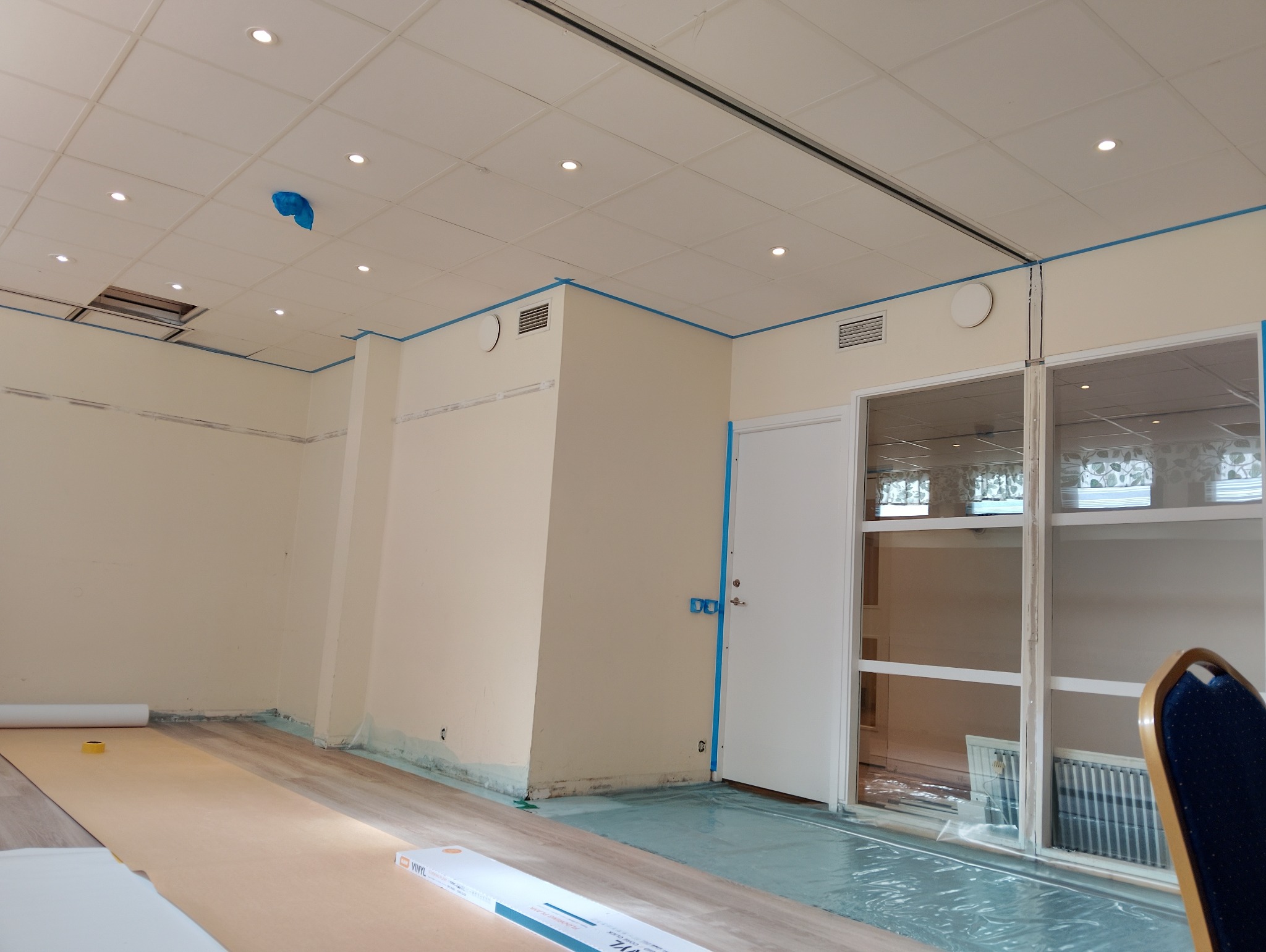 Wnętrze w trakcie układania paneli podłogowych. Podłoga częściowo zakryta folią ochronną i kartonem. Ściany pomalowane, zabezpieczone taśmą malarską. Widoczne puszki paneli.