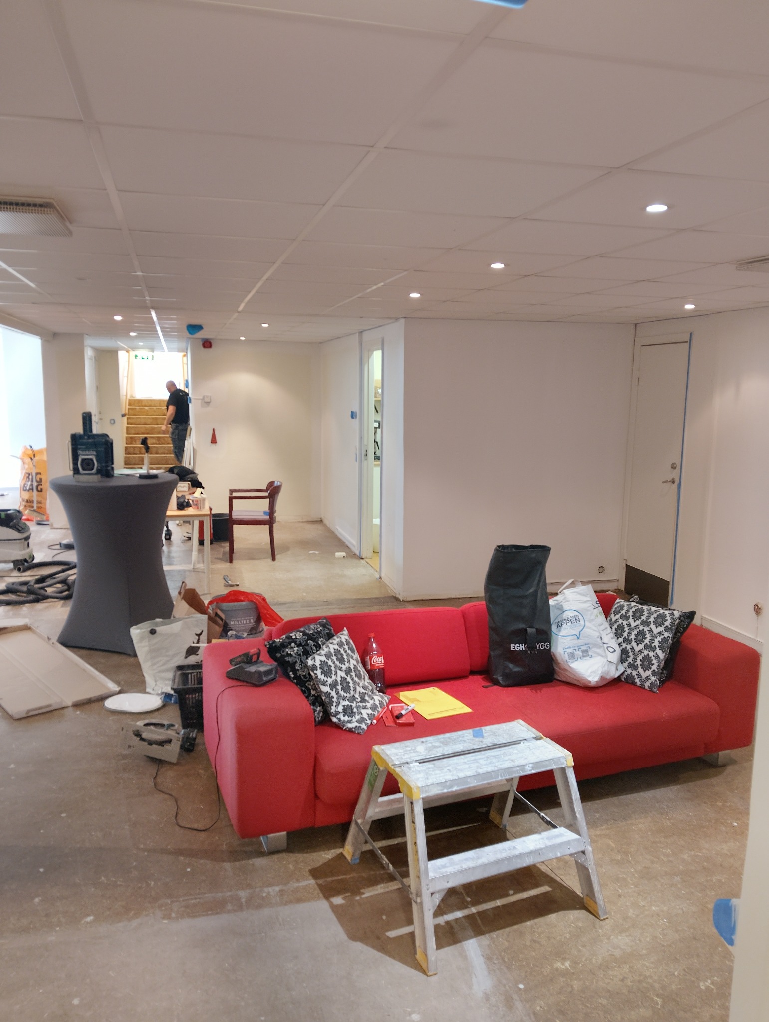 Pokój w trakcie remontu: czerwona sofa z poduszkami, drabina, narzędzia i worki budowlane na podłodze. W tle widoczna osoba na schodach. Oświetlenie sufitowe.