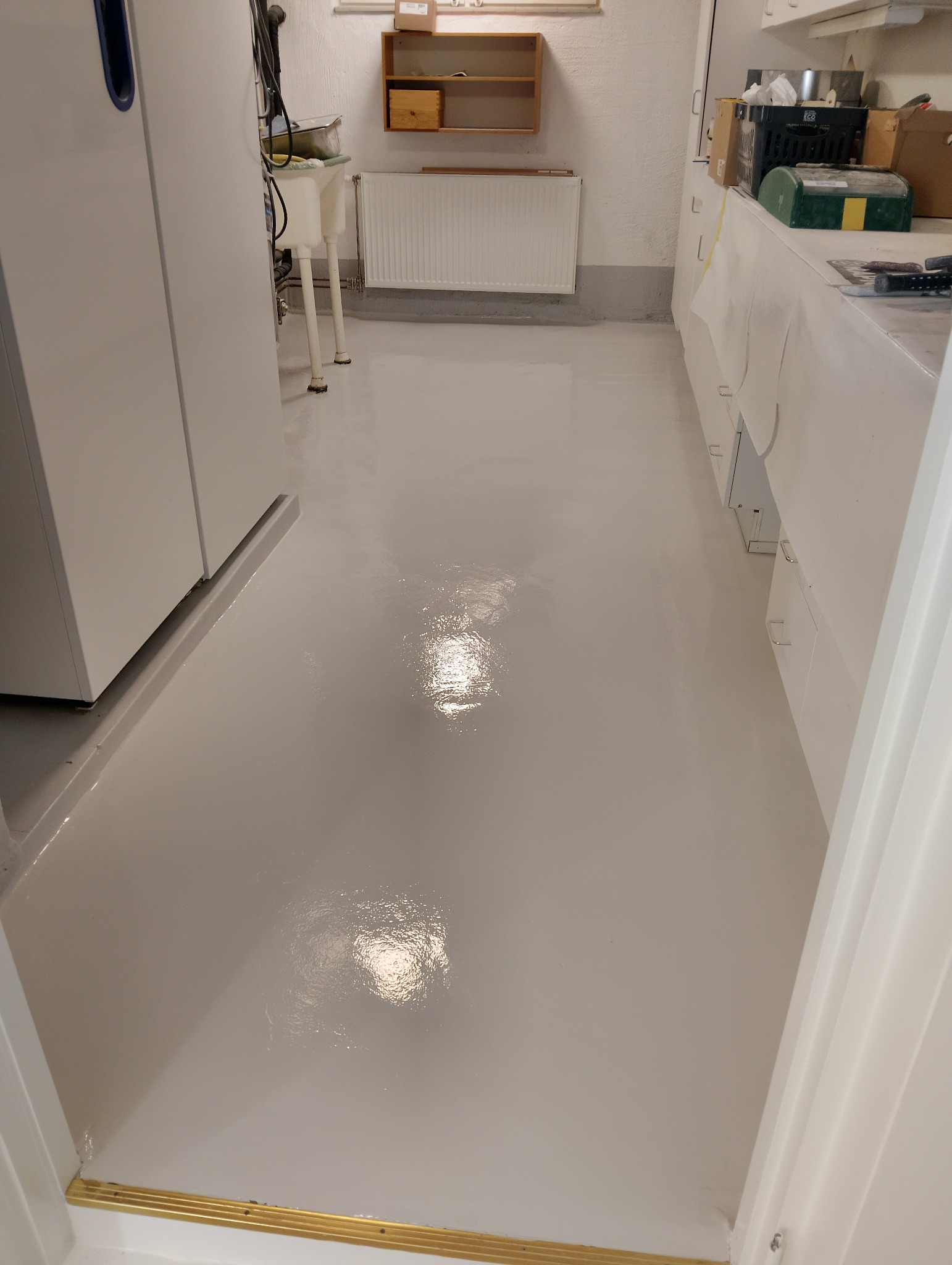 Błyszcząca, jasnoszara posadzka z żywicy epoksydowej w pomieszczeniu gospodarczym. Widoczne białe szafki i elementy instalacji. Podłoga odbija światło.
