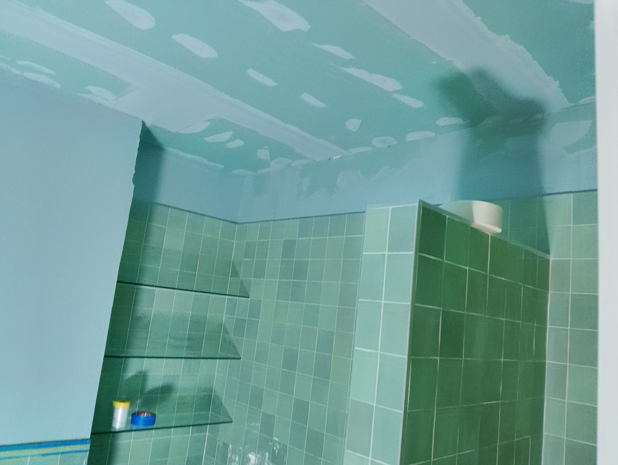 Niebiesko-zielona łazienka w trakcie remontu. Sufit i ściany pokryte wstępną warstwą gładzi, widoczne płytki na ścianach. Na półce stoją pojemniki z farbą.