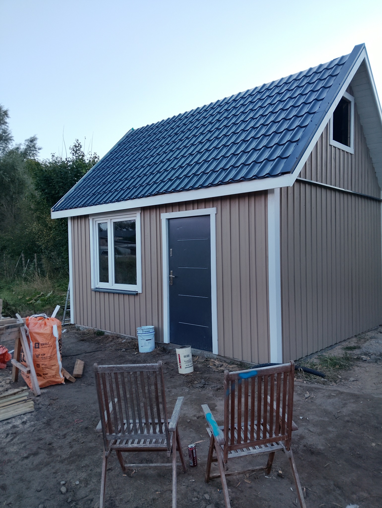 Mały, parterowy budynek z ciemnym dachem i beżową elewacją z pionowych paneli. Widoczne okno, drzwi wejściowe i dwa krzesła na zewnątrz.