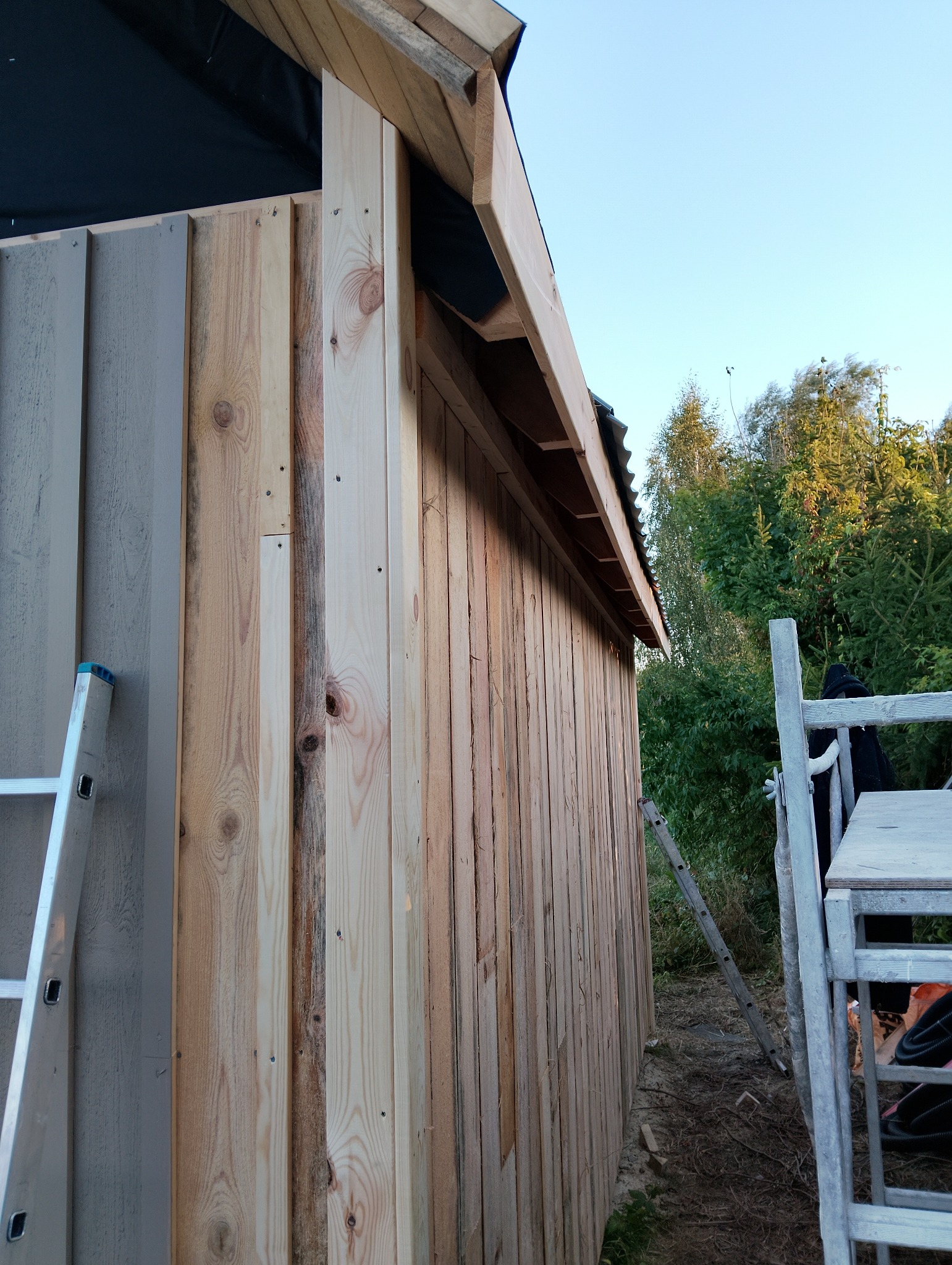 Budowa drewnianej konstrukcji z widocznymi deskami i drabinkami, częściowo pomalowana na szaro, w tle zieleń drzew.