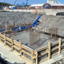 Szary, betonowy fundament w drewnianym szalunku z wystającymi prętami zbrojeniowymi na placu budowy. W tle budynek Renta i sprzęt budowlany.