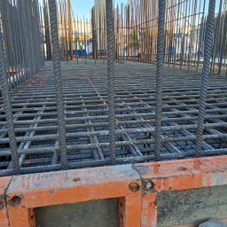 Artsteel - Zbrojenie fundamentów z widocznymi prętami pionowymi i poziomymi tworzącymi siatkę, częściowo zalane betonem, z pomarańczowymi elementami szalunku.