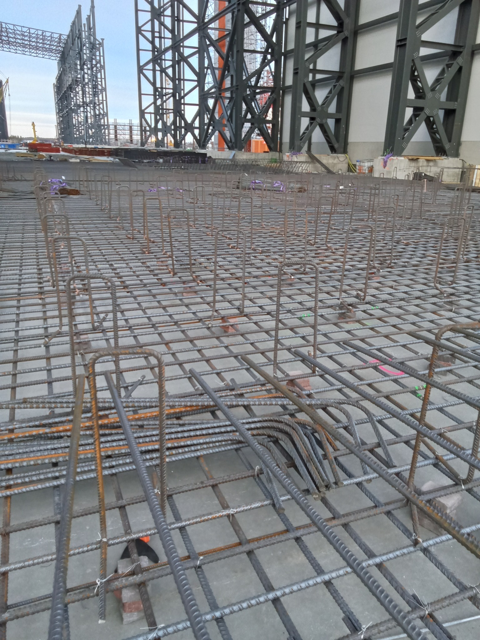 Siatka zbrojeniowa na budowie hali, widoczne stalowe konstrukcje w tle i betonowa podstawa. Detal zbrojenia z bliska, etap prac fundamentowych.
