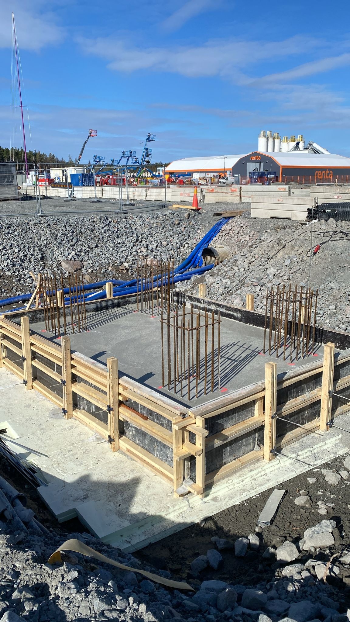 Szary, betonowy fundament w drewnianym szalunku z wystającymi prętami zbrojeniowymi na placu budowy. W tle budynek Renta i sprzęt budowlany.