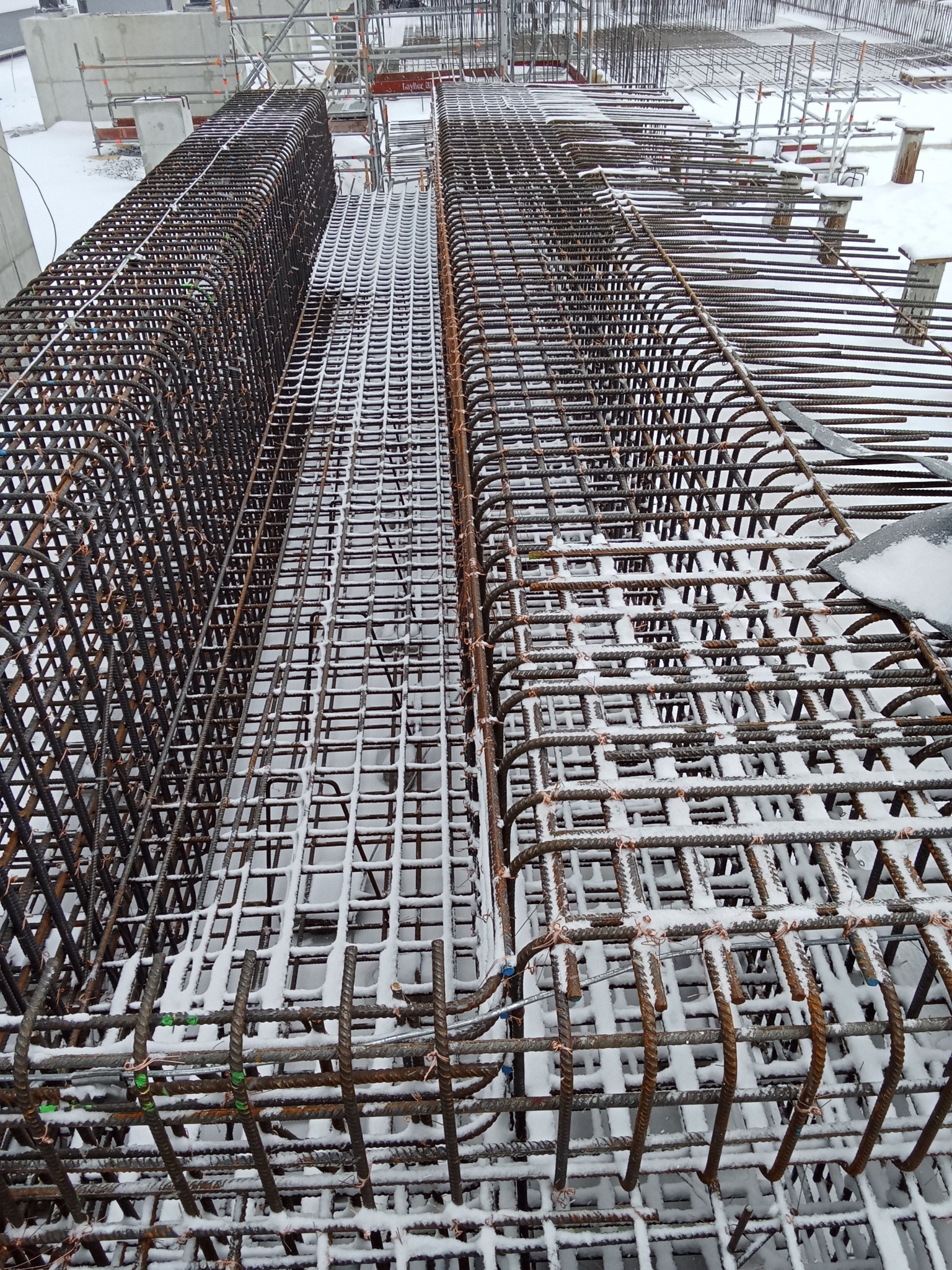 Zbrojenie w szalunku, pokryte śniegiem na budowie. Widok z góry ukazuje siatkę prętów i detale konstrukcji, przygotowane do zalania betonem w zimowych warunkach.