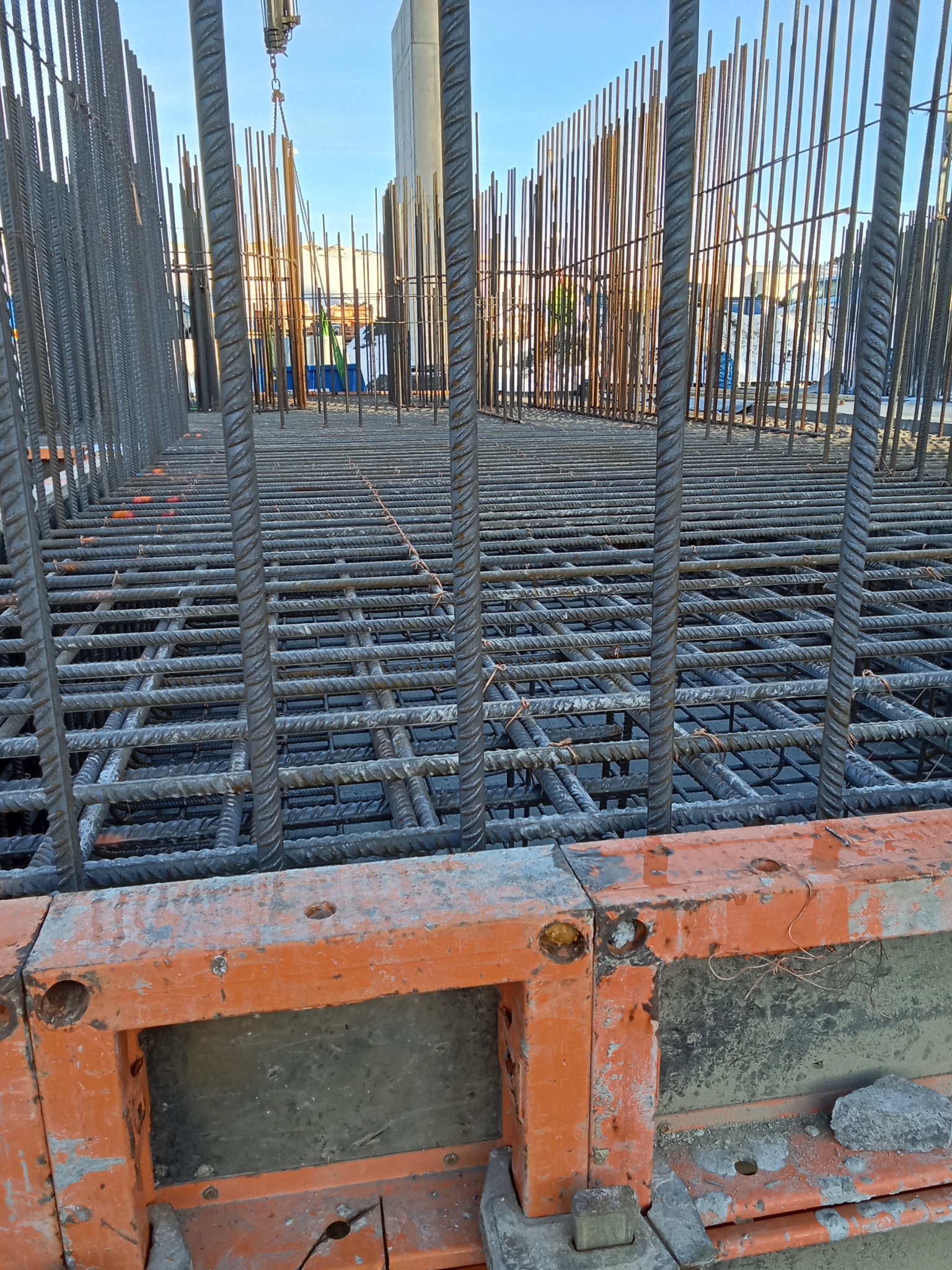 Zbrojenie fundamentów z widocznymi prętami pionowymi i poziomymi tworzącymi siatkę, częściowo zalane betonem, z pomarańczowymi elementami szalunku.