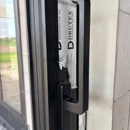 Drutex Susz - Czarne okno PCV Drutex z naklejką ochronną i uchwytem, zamontowane w surowej ścianie. Widoczne detale wykończenia i częściowo usunięta folia zabezpieczająca.