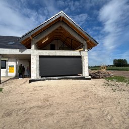 LUX BUD SPÓŁKA Z OGRANICZONĄ ODPOWIEDZIALNOŚCIĄ - Ciemnoszara brama garażowa segmentowa w surowym budynku z betonu komórkowego, widok z zewnątrz, w tle błękitne niebo i osoby przy tablicy informacyjnej.