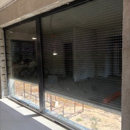 LUX BUD SPÓŁKA Z OGRANICZONĄ ODPOWIEDZIALNOŚCIĄ - Zamknięta roleta zewnętrzna w oknie z czarną ramą, widok z wnętrza surowego pomieszczenia. Roleta częściowo opuszczona, widoczne szczeliny. Na zewnątrz widok na plac budowy.