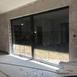 LUX BUD SPÓŁKA Z OGRANICZONĄ ODPOWIEDZIALNOŚCIĄ - Zamknięta roleta zewnętrzna w oknie z czarną ramą, widok z wnętrza surowego pomieszczenia w trakcie wykończenia. Na podłodze resztki materiałów budowlanych.