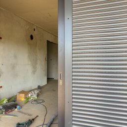 LUX BUD SPÓŁKA Z OGRANICZONĄ ODPOWIEDZIALNOŚCIĄ - Zamknięta, szara roleta wewnętrzna w surowym wnętrzu. Widoczne narzędzia i materiały budowlane na podłodze. W tle ściany w stanie deweloperskim.