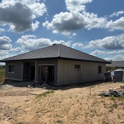 LUX BUD SPÓŁKA Z OGRANICZONĄ ODPOWIEDZIALNOŚCIĄ - Surowy dom jednorodzinny z ciemną dachówką, widoczne okna z roletami zewnętrznymi, teren wokół w trakcie prac wykończeniowych, błękitne niebo z chmurami.