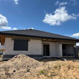 LUX BUD SPÓŁKA Z OGRANICZONĄ ODPOWIEDZIALNOŚCIĄ - Dom w trakcie budowy z zamontowaną roletą zewnętrzną. Widoczny surowy stan ścian z betonu komórkowego, czarna dachówka i sterta piasku na pierwszym planie.