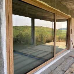 LUX BUD SPÓŁKA Z OGRANICZONĄ ODPOWIEDZIALNOŚCIĄ - Duże przesuwne okno z drewnianą ramą w surowym stanie budynku, z widokiem na zielony krajobraz. Widoczne detale montażu i surowy beton.