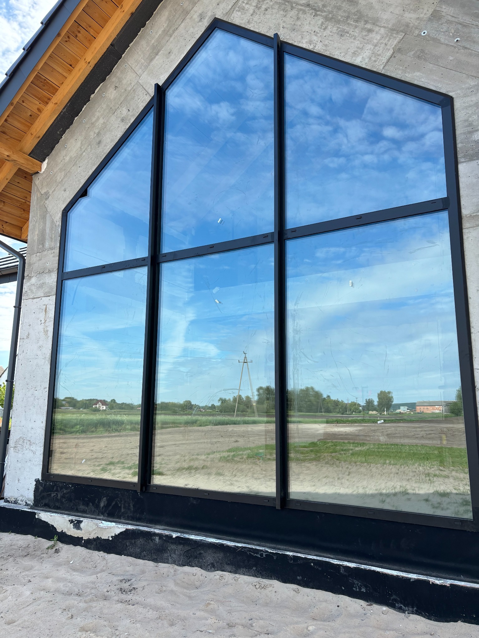 Duże, trapezowe okno z czarną ramą w surowym, betonowym budynku w trakcie budowy. W szybach odbija się błękitne niebo i krajobraz.