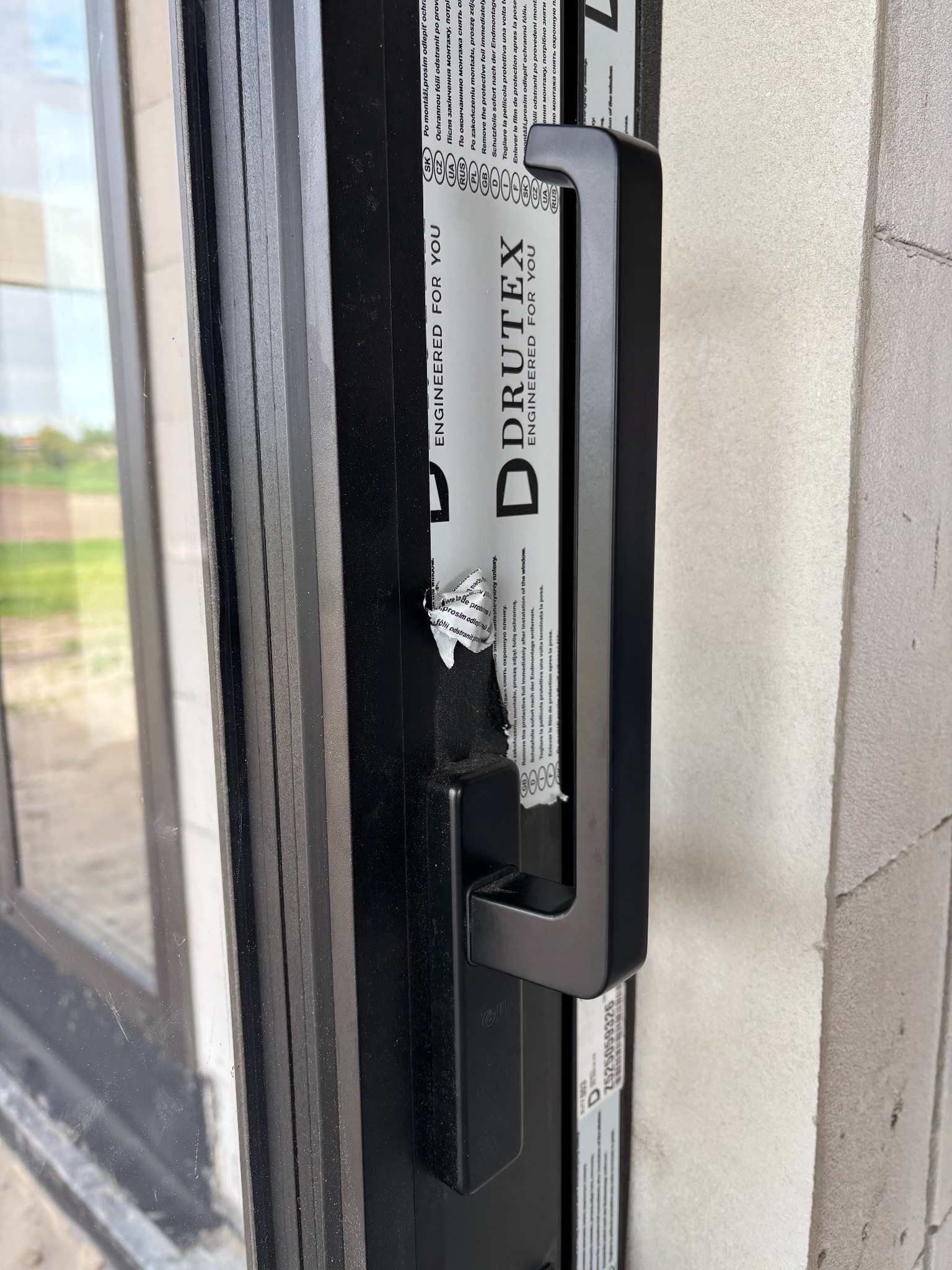 Czarne okno PCV Drutex z naklejką ochronną i uchwytem, zamontowane w surowej ścianie. Widoczne detale wykończenia i częściowo usunięta folia zabezpieczająca.