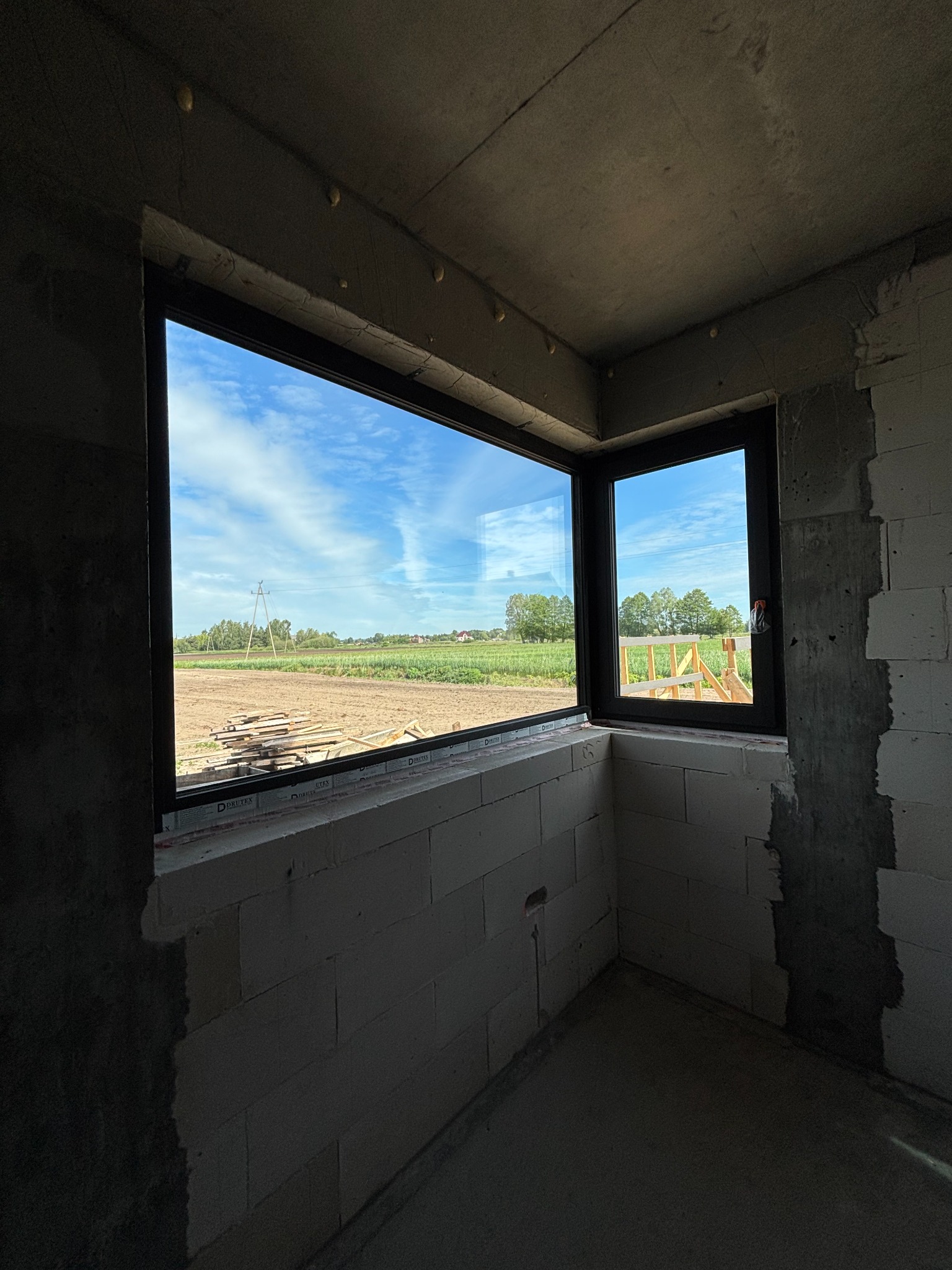 Czarne ramy okienne w surowym wnętrzu budynku w Suszu, z widokiem na pole i błękitne niebo. Okna zamontowane w ścianie z białych bloczków.