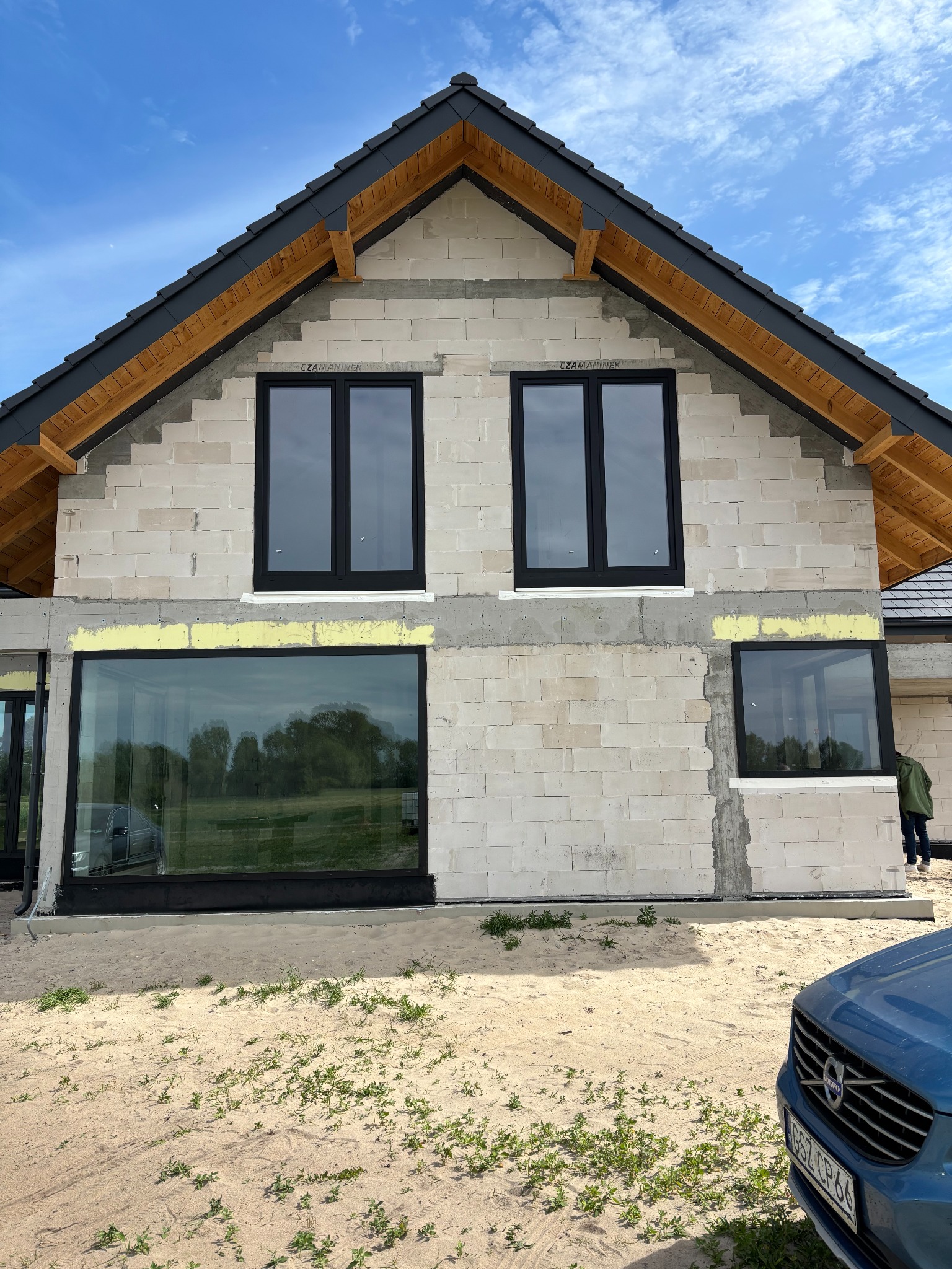 Dom w trakcie budowy z zamontowanymi oknami w czarnych ramach. Widoczne bloczki z betonu komórkowego i drewniana konstrukcja dachu. Na pierwszym planie fragment auta.