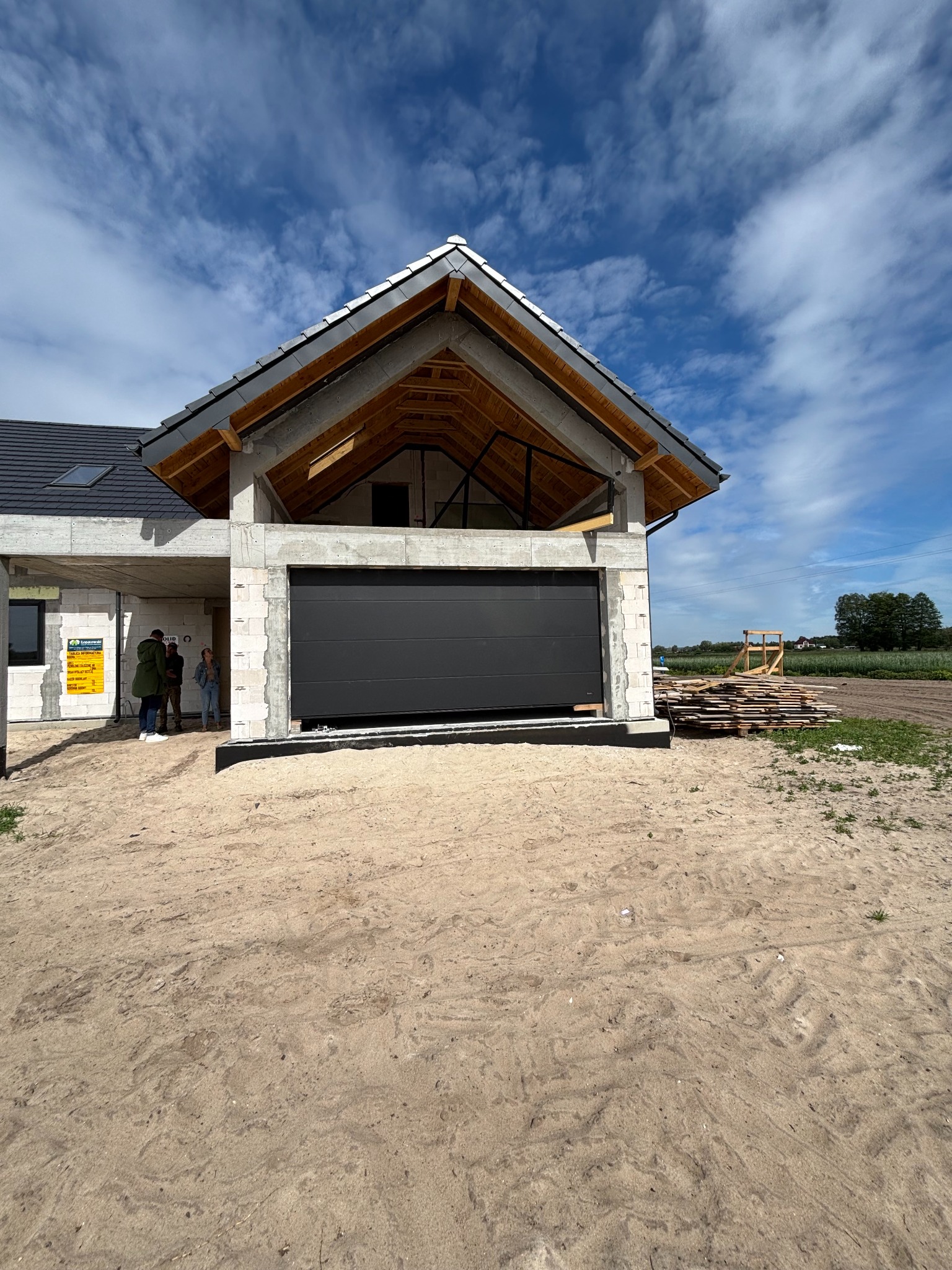 Ciemnoszara brama garażowa segmentowa w surowym budynku z betonu komórkowego, widok z zewnątrz, w tle błękitne niebo i osoby przy tablicy informacyjnej.