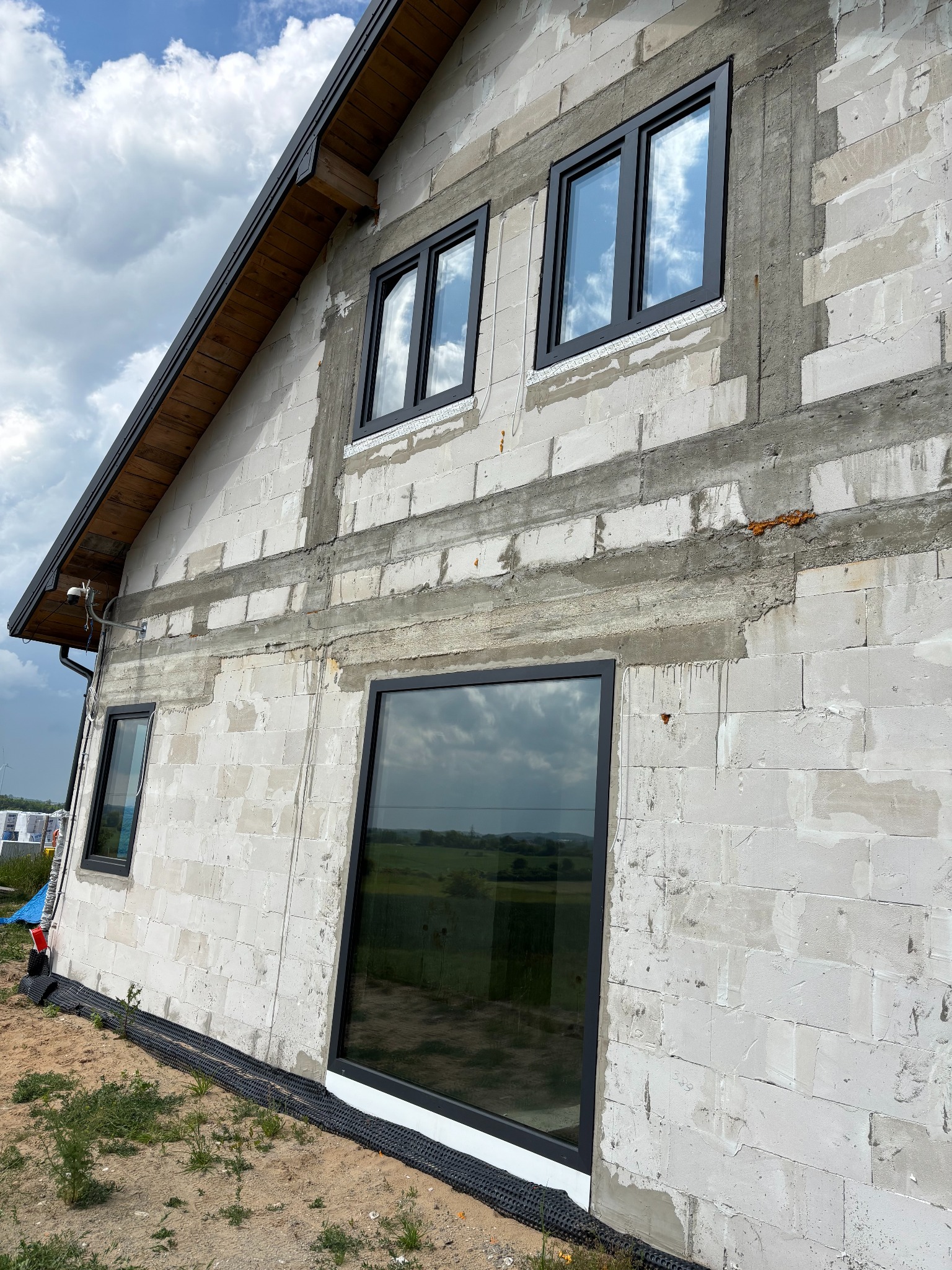 Surowy budynek z zamontowanymi oknami w czarnych ramach, odbijającymi niebo. Widoczne bloczki i zaprawa, teren wokół budynku niezagospodarowany.