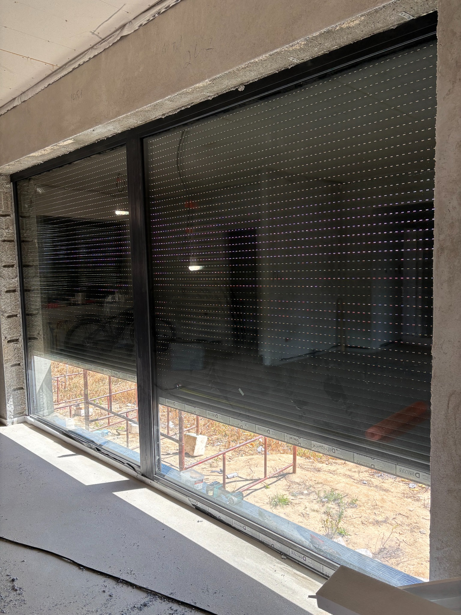 Zamknięta roleta zewnętrzna w oknie z czarną ramą, widok z wnętrza surowego pomieszczenia. Roleta częściowo opuszczona, widoczne szczeliny. Na zewnątrz widok na plac budowy.